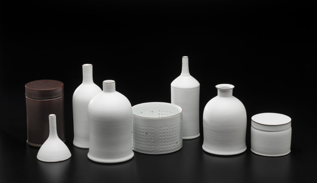 A row of eight small bottles and containers, all in white porcelain except one brown cylindrical box on far left. All photographed against black background.